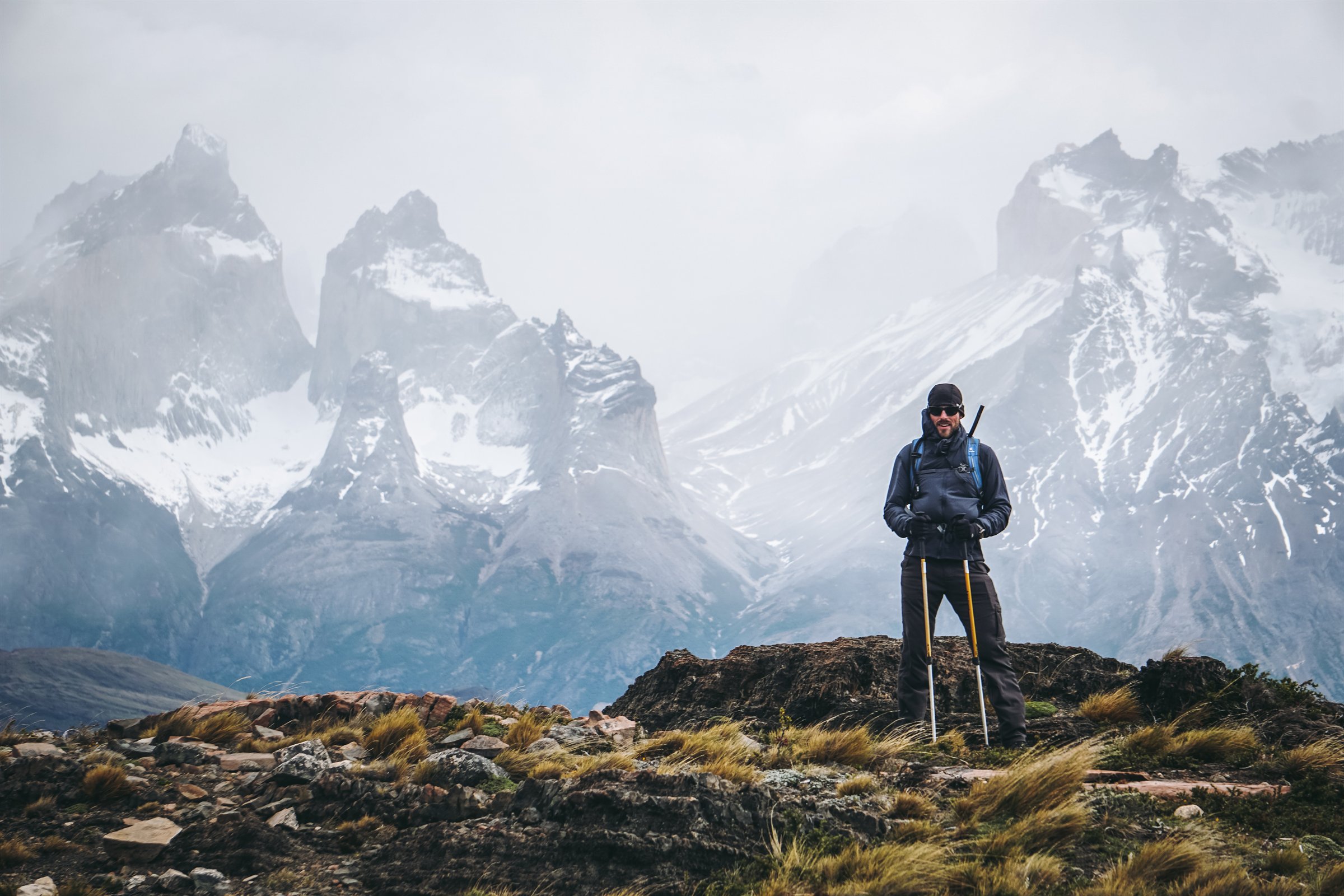 TJ standing with trekking poles in front of a wide, snow-covered mountain range
